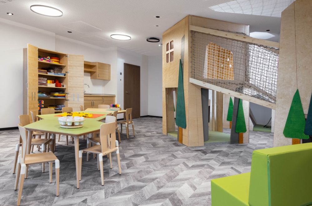 Lichtdurchfluteter Aufenthaltsraum mit Klettergerüst für Kinder sowie mehreren niedrigen Tischen und Stühlen.