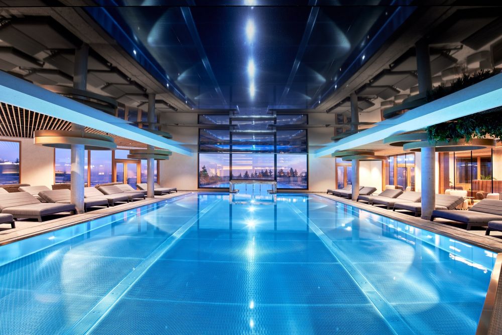 Blick auf den hellblauen Indoorpool mit Liegen, im Hintergrund der Outdoorpool.