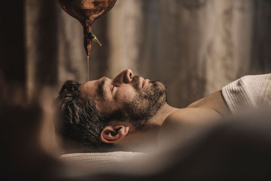 Nahaufnahme eines liegenden Mannes mit geschlossenen Augen, dem Öl auf seine Stirn läuft.