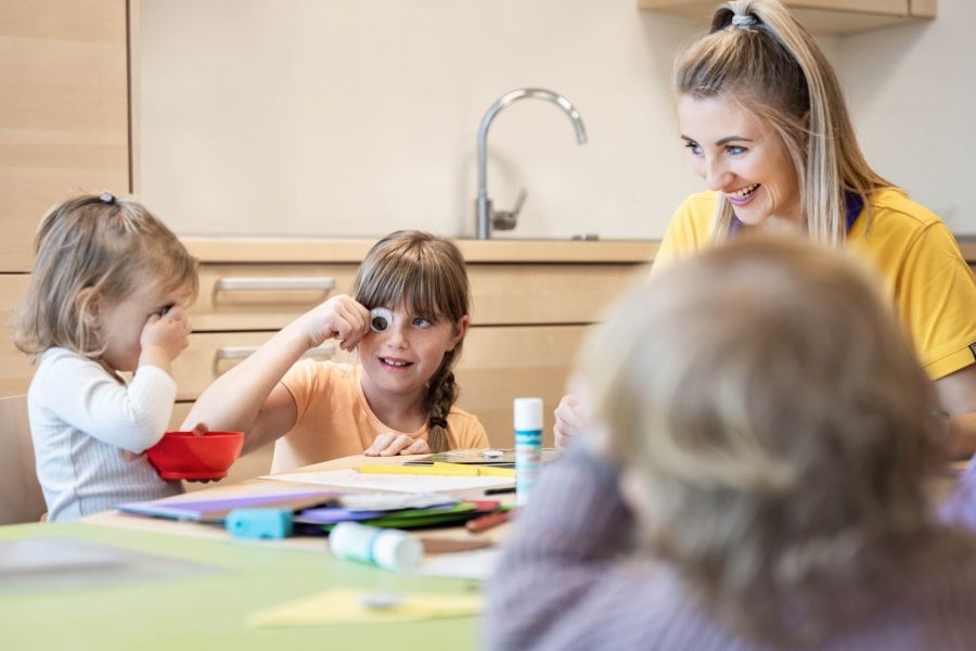 Drei Kinder sitzen mit einer jungen Frau an einem niedrigen Tisch und basteln. Drei Kinder sitzen mit einer jungen Frau an einem niedrigen Tisch und basteln.